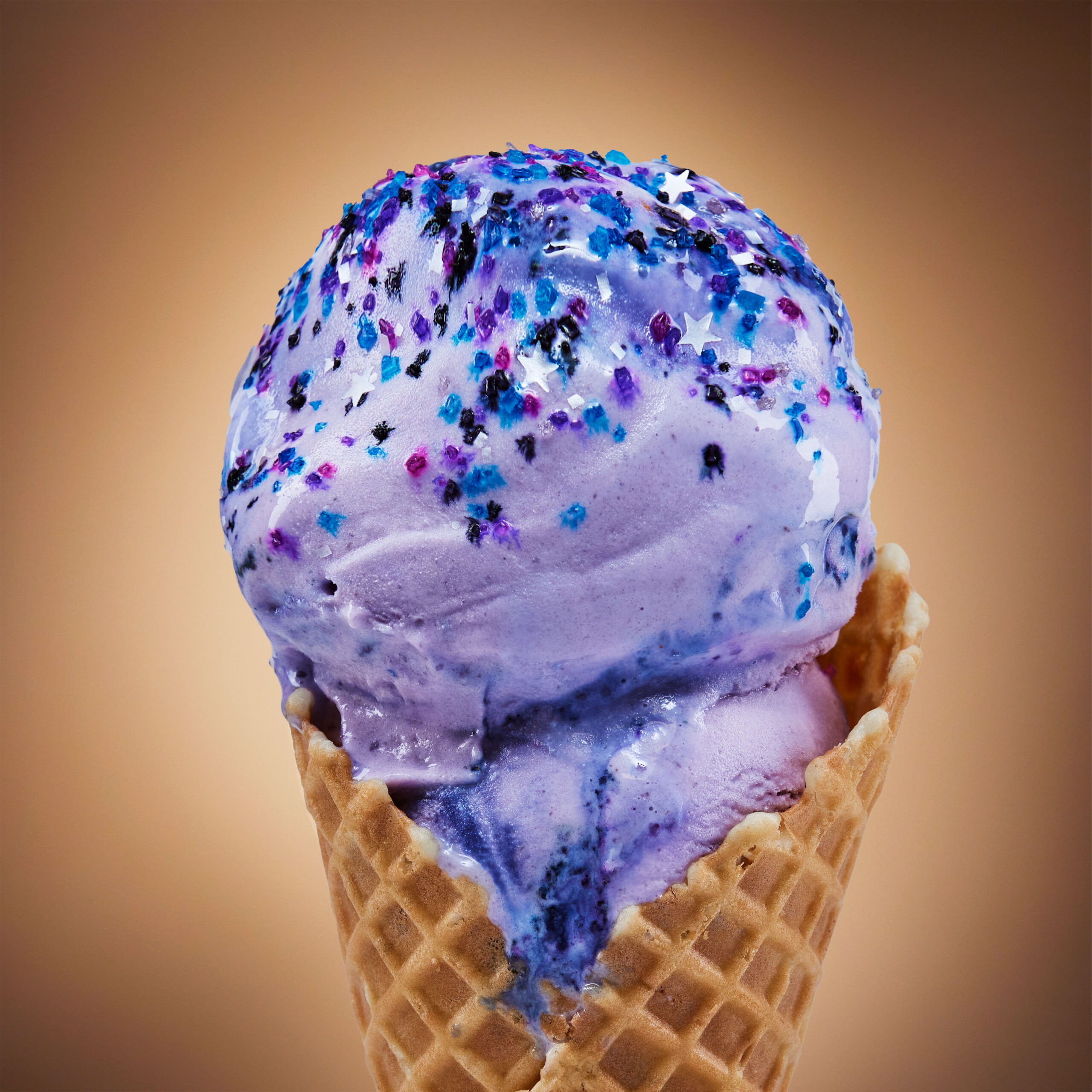 A double scoop of purple ice cream with silver black magenta and blue star sprinkles on a waffle cone