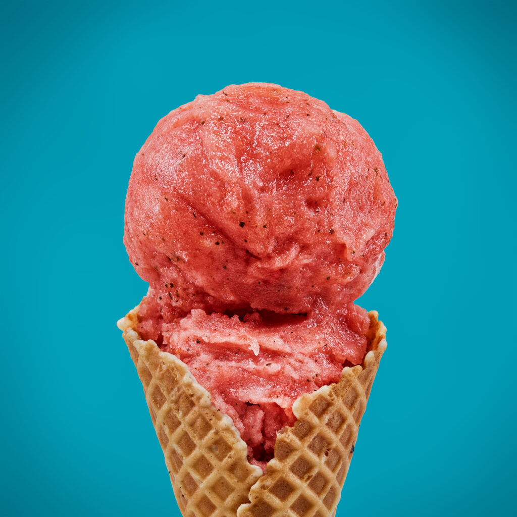 a double scoop of pink sorbet with flecks of green mint leaves, in a waffle cone, against a bright blue back ground